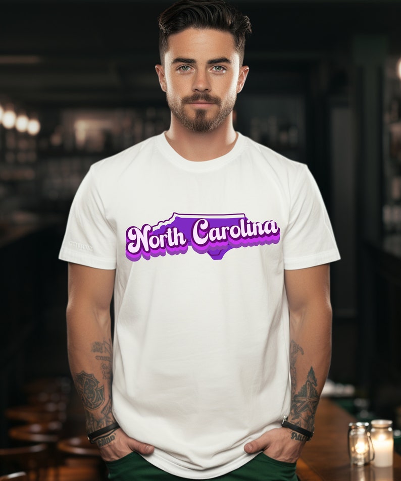 Może przedstawiać: Biała koszulka z fioletowym obrysem stanu Karolina P&oacute;łnocna i tekstem "North Carolina" w retro czcionce.