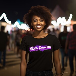 Może przedstawiać: Kobieta w czarnym t-shircie z fioletowym nadrukiem przedstawiającym stan Karolina P&oacute;łnocna i tekstem "North Carolina".
