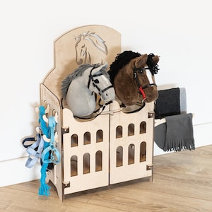 Op de afbeelding: Een houten speelgoedpaardenstal met twee stokpaardenkoppen, één grijze en één bruine. De stal heeft een decoratieve paardenkopuitsparing en boogvormige openingen. Blauwe en witte speelgoedpaardentuig hangt aan de zijkant. Een grijze deken is over de zijkant gedrapeerd.
