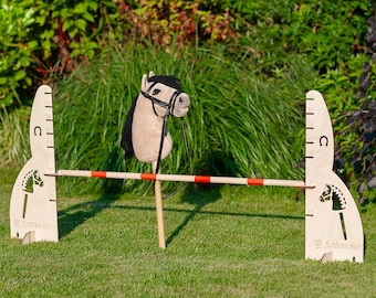 May include: A wooden horse jumping obstacle with a red and white bar. The obstacle is made of two wooden panels with a horse head cutout and a measurement scale. The horse head is a plush toy with a black bridle.