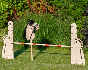 May include: A wooden horse jumping over a red and white bar. The horse is made of wood and has a brown and black mane and tail. The bar is attached to two wooden posts with a horse head design. The posts have a ruler on them, with measurements in centimeters. The background is a green grassy lawn.