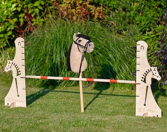 May include: A wooden horse jumping obstacle with a height adjustable feature. The obstacle is made of wood and has a red and white stripe. The horse is a stuffed toy with a black bridle and a brown head. The obstacle is set up on a green grassy lawn.