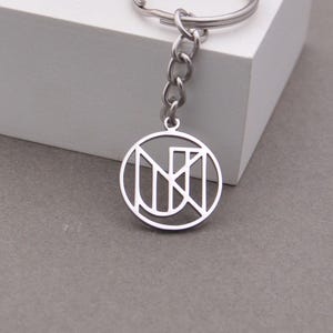 Może przedstawiać: Srebrny brelok do kluczy z okrągłą zawieszką z abstrakcyjnym wzorem. Zawieszka jest połączona z łańcuszkiem i kółkiem. Brelok jest wykonany z metalu i jest prezentowany na białej powierzchni na szarym tle.