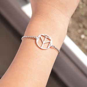Puede incluir: Una pulsera plateada con un colgante circular geométrico. El colgante tiene un diseño recortado con formas angulares. La pulsera está en una cadena y se lleva en la muñeca.
