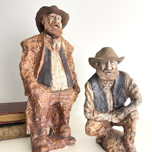 May include: Two ceramic figurines of elderly men. One stands, the other kneels. Both wear hats, vests, and jackets in shades of brown and tan. The figures have detailed facial features and textured clothing, suggesting a rustic or folk art style.