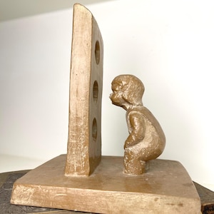 May include: A tan-colored bookend featuring a rectangular panel with three circular cutouts, and a small figure in a squatting position. The bookend is made of a textured material and sits on a rectangular base.