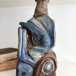 May include: A sculptural ceramic bottle with a glossy finish, featuring a blend of deep blue, brown, and tan glazes. The object has an irregular, abstract form with a narrow neck and a textured, layered surface that transitions from dark brown at the base.