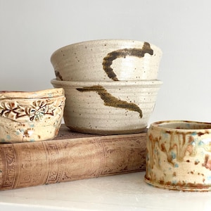 May include: A collection of handmade ceramic bowls and a small pot. The bowls are beige with brown abstract designs. The small pot has a floral design and the other has a dripped glaze. The items are on a vintage book.