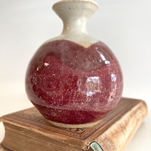 May include: A small, round ceramic vase with a burgundy body and a cream-colored neck. The vase is sitting on top of a vintage book with a worn, brown cover. The vase has a glossy finish.