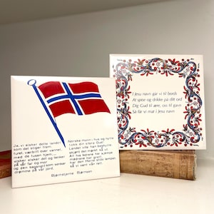 May include: Two square white ceramic tiles featuring Norwegian cultural themes. One tile displays a red, white, and blue Norwegian flag with the text of the national anthem by Bjørnstjerne Bjørnson. The other tile features a floral border and prayer text.