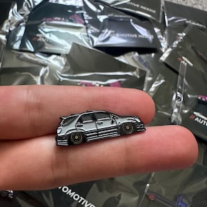 May include: A silver enamel pin of a silver car with black accents. The pin is on a finger. The pin is part of a collection of pins with the text "Automotive Needz" on the packaging.