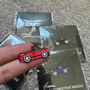 May include: A set of four enamel pins featuring different colored pickup trucks. The pins are all the same design, but each one is a different color: red, black, white, and blue. The pins are all on a black background with the text "//AUTOMOTIVE NEEDZ" printed on the bottom.