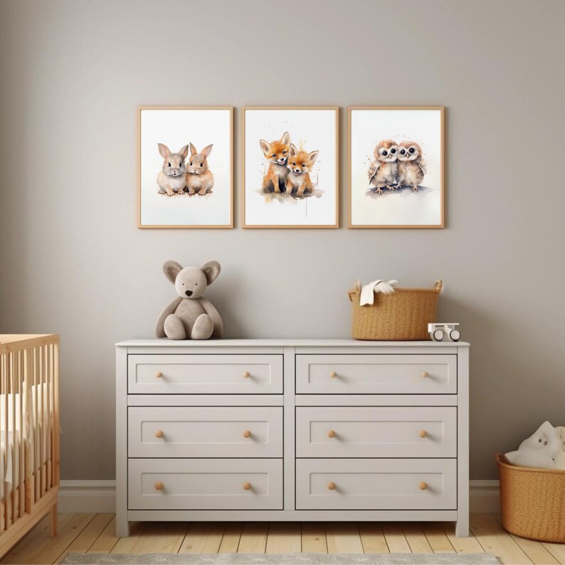 May include: Three framed watercolor paintings of animal pairs. The first painting shows two bunnies, the second shows two foxes, and the third shows two owls. The paintings are hung on a light gray wall above a white dresser with six drawers.