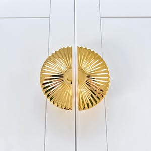 May include: A pair of gold-toned cabinet door pulls with a sunburst design. The pulls are split in half and are mounted on a white cabinet door.