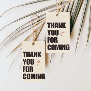 May include: Two brown paper gift tags with the text "Thank You For Coming" in black lettering and red stars. The tags are hanging from twine against a white background with a palm leaf.