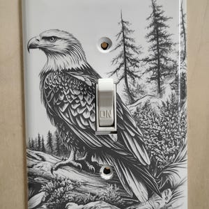 May include: White light switch cover featuring a detailed black and white illustration of a bald eagle perched on a log with a forest backdrop. The switch is white, with "ON" printed on it.