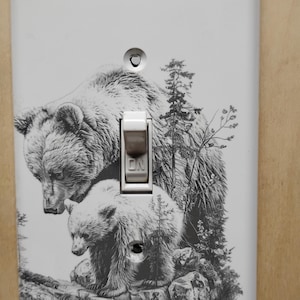 May include: White light switch cover featuring a detailed black and white illustration of two bears in a forest setting. The switch is in the "ON" position. The design includes trees and rocky terrain, creating a nature-themed aesthetic.