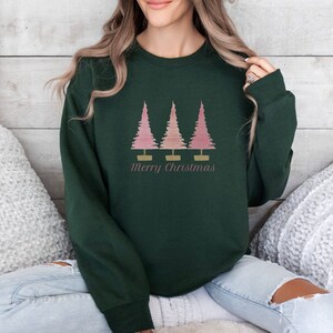 May include: Dark green sweatshirt featuring a graphic of three pink Christmas trees with gold bases. The text "Merry Christmas" is printed below the trees in a script font. The sweatshirt is a long-sleeved crew neck.