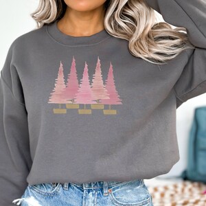 May include: A dark gray sweatshirt featuring a pink and gold tree design. The graphic shows five stylized trees in varying shades of pink, with gold rectangular bases. The sweatshirt has a crew neck and long sleeves.