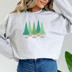 May include: A light gray sweatshirt featuring a graphic of five stylized green Christmas trees. The trees vary in shades of green and are set on a light brown base. The sweatshirt has a crew neck and long sleeves.