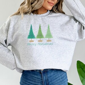 May include: A light gray sweatshirt featuring three stylized Christmas trees in shades of green. The trees are set on brown bases, with the words "Merry Christmas" printed below in a matching green color. The sweatshirt is a casual, festive garment.