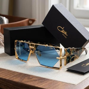 May include: Gold-tone metal sunglasses with geometric blue lenses and tortoiseshell-patterned temples. The frames feature the Cazal logo on the lens and temple. Includes a black branded box and a small black informational card with the Cazal logo.