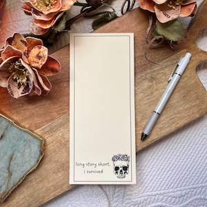 May include: A white notepad with a black border and a skull with a floral crown at the bottom. The notepad has the text "long story short, i survived" printed on it.