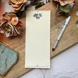 May include: A white notepad with a bee and honeycomb design on the top. The notepad is on a wooden surface with a pen next to it.