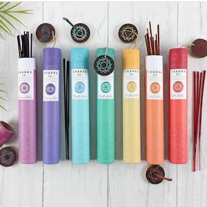 Peut inclure: Sept bâtonnets d'encens colorés avec des symboles et des noms de chakras. Les bâtonnets sont emballés dans des tubes individuels avec un symbole de chakra en bois sur le dessus. Les tubes sont disposés en rangée sur une surface en bois blanc.