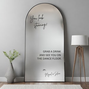 May include: A large, arched mirror with a black frame. The mirror has text that reads "You look stunning!" and "GRAB A DRINK AND SEE YOU ON THE DANCE FLOOR." A floor lamp and a vase with greenery are visible.