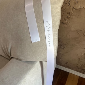 May include: A beige cushion with two white satin ribbons attached. The ribbons have the word "Matchhunter" printed in a cursive font. The cushion is on a sofa.
