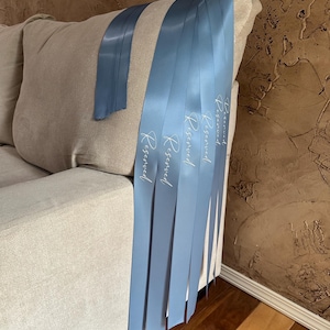Peut inclure: Plusieurs rubans de satin bleu clair avec le mot "Reserved" imprim&eacute; en blanc sont drap&eacute;s sur le bras d'un canap&eacute; beige. Un ruban est s&eacute;par&eacute;. Les rubans sont d'un bleu clair.