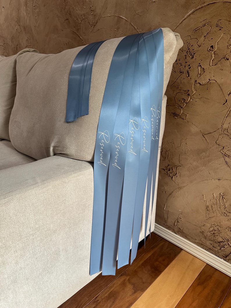 Peut inclure: Plusieurs rubans de satin bleu clair avec le mot "Reserved" imprim&eacute; en blanc sont drap&eacute;s sur le bras d'un canap&eacute; beige. Un ruban est s&eacute;par&eacute;. Les rubans sont d'un bleu clair.