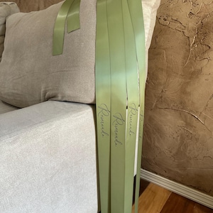 Peut inclure: Gros plan sur des rubans de satin vert clair avec le mot "Reservada" imprim&eacute; en &eacute;criture noire. Les rubans sont drap&eacute;s sur un coussin de canap&eacute; beige. L'arri&egrave;re-plan est un mur brun textur&eacute;.