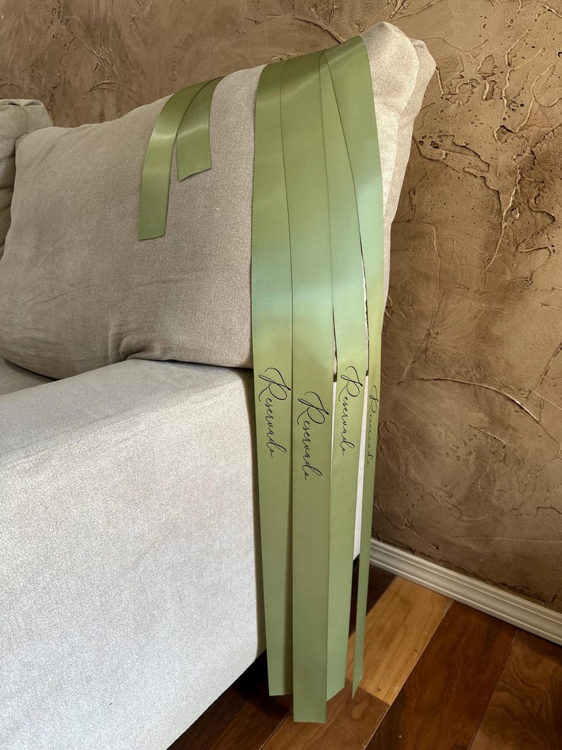 Peut inclure: Gros plan sur des rubans de satin vert clair avec le mot "Reservada" imprim&eacute; en &eacute;criture noire. Les rubans sont drap&eacute;s sur un coussin de canap&eacute; beige. L'arri&egrave;re-plan est un mur brun textur&eacute;.