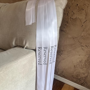 Peut inclure: Rubans de satin blancs avec le mot "Reserved" imprim&eacute; en noir. Les rubans sont drap&eacute;s sur un canap&eacute; beige. L'arri&egrave;re-plan pr&eacute;sente un mur brun textur&eacute;.
