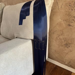 Peut inclure: Rubans de satin bleu marine avec le mot "Reserved" en script dor&eacute; drap&eacute;s sur un coussin de canap&eacute; beige. Les rubans sont longs et pendent, cr&eacute;ant un accent d&eacute;coratif. Le fond pr&eacute;sente un mur brun textur&eacute; et un sol en bois.