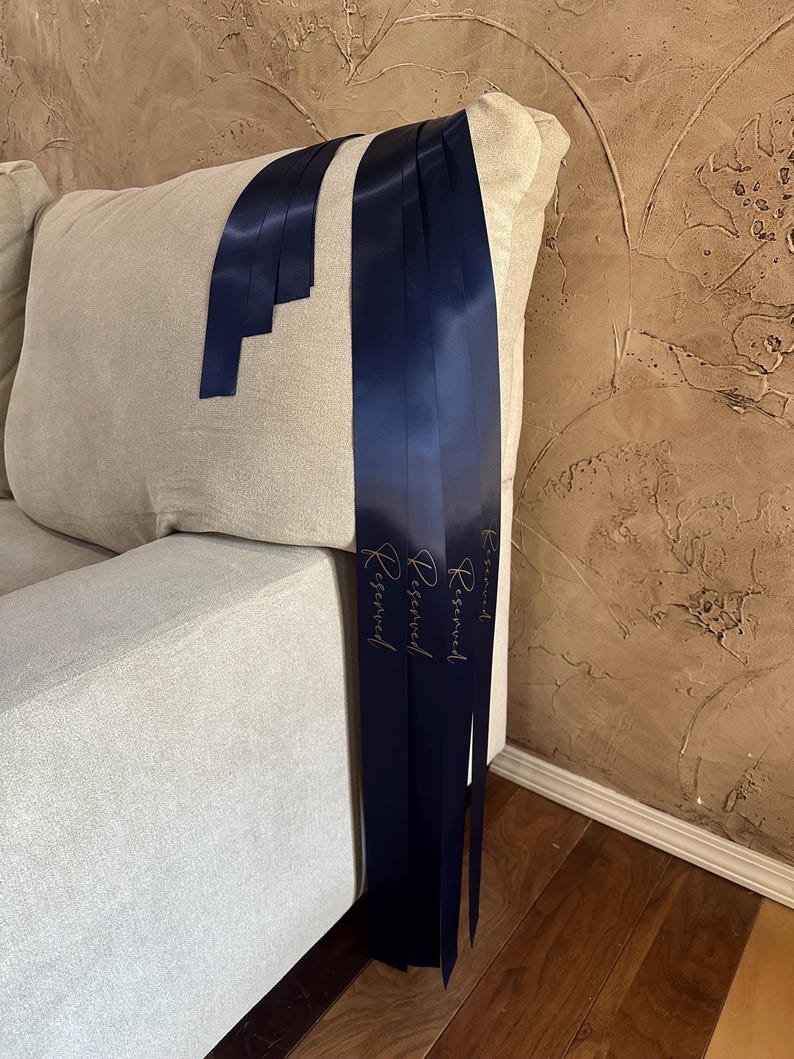 Peut inclure: Rubans de satin bleu marine avec le mot "Reserved" en script dor&eacute; drap&eacute;s sur un coussin de canap&eacute; beige. Les rubans sont longs et pendent, cr&eacute;ant un accent d&eacute;coratif. Le fond pr&eacute;sente un mur brun textur&eacute; et un sol en bois.