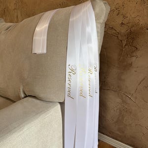 Peut inclure: Rubans de satin blancs avec le mot "Reserved" en lettres dor&eacute;es drap&eacute;s sur le bras d'un canap&eacute; beige. Les rubans sont pli&eacute;s et pendent, cr&eacute;ant un accent d&eacute;coratif. L'arri&egrave;re-plan est un mur brun textur&eacute;.