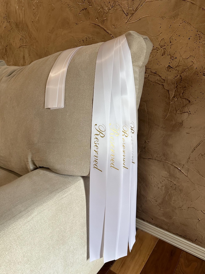 Peut inclure: Rubans de satin blancs avec le mot "Reserved" en lettres dor&eacute;es drap&eacute;s sur le bras d'un canap&eacute; beige. Les rubans sont pli&eacute;s et pendent, cr&eacute;ant un accent d&eacute;coratif. L'arri&egrave;re-plan est un mur brun textur&eacute;.