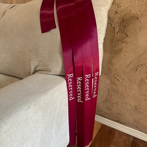 Peut inclure: Rubans de satin bordeaux avec le mot "Reserved" imprim&eacute; en blanc. Les rubans sont drap&eacute;s sur le bras d'un canap&eacute; beige. L'arri&egrave;re-plan pr&eacute;sente un mur textur&eacute; et un sol en bois.
