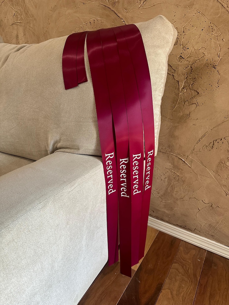 Peut inclure: Rubans de satin bordeaux avec le mot "Reserved" imprim&eacute; en blanc. Les rubans sont drap&eacute;s sur le bras d'un canap&eacute; beige. L'arri&egrave;re-plan pr&eacute;sente un mur textur&eacute; et un sol en bois.