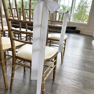May include: Gold-colored wooden chairs with cream-colored seats are arranged in rows. Each chair is adorned with a white ribbon tied in a bow, with the word "Reserved" printed on the ribbon. The chairs are set in a well-lit room with large windows.