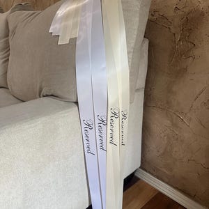 Peut inclure: Trois rubans de satin blancs, ivoire et cr&egrave;me, chacun imprim&eacute; du mot "Reserved" en &eacute;criture &eacute;l&eacute;gante. Les rubans sont drap&eacute;s sur le bras d'un canap&eacute; gris clair, avec les extr&eacute;mit&eacute;s visibles.
