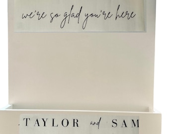 May include: A white rectangular wooden wall sign featuring a built-in storage pocket at the bottom. The top section displays the text WELCOME we're so glad you're here in black. The pocket displays the text TAYLOR and SAM MARCH 17, 2024 in black.