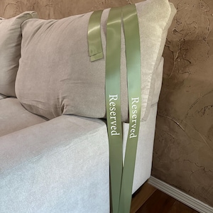 Peut inclure: Un canap&eacute; gris clair avec un grand coussin moelleux. Deux rubans vert olive avec le mot "Reserved" imprim&eacute; en blanc sont drap&eacute;s sur le canap&eacute;. L'arri&egrave;re-plan est un mur textur&eacute; et brun.