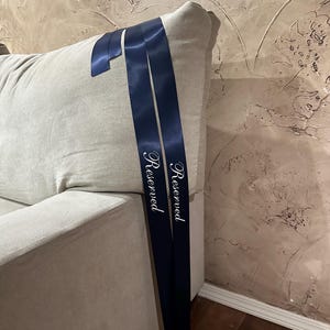 Peut inclure: Un canap&eacute; gris clair avec un ruban bleu marine drap&eacute; sur le dossier. Le ruban porte le mot "Reserved" imprim&eacute; en &eacute;criture blanche. Le canap&eacute; est contre un mur textur&eacute; brun clair.