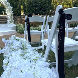 May include: White folding chairs with black ribbons that read "Reserved" are set up for an outdoor event. A pathway of white flowers leads to a floral arch. The scene is set on a grassy area with green hedges in the background.