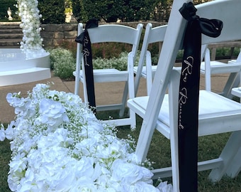 May include: White folding chairs with black ribbons that read "Reserved" are set up for an outdoor event. A pathway of white flowers leads to a floral arch. The scene is set on a grassy area with green hedges in the background.