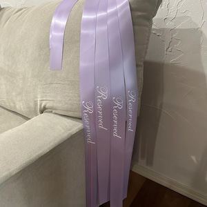 Peut inclure: Plusieurs rubans de satin lavande avec le mot &laquo;&nbsp;Reserved&nbsp;&raquo; imprim&eacute; en &eacute;criture blanche sont drap&eacute;s sur un canap&eacute; beige. Les rubans sont dispos&eacute;s en cascade, avec les extr&eacute;mit&eacute;s des rubans visibles.